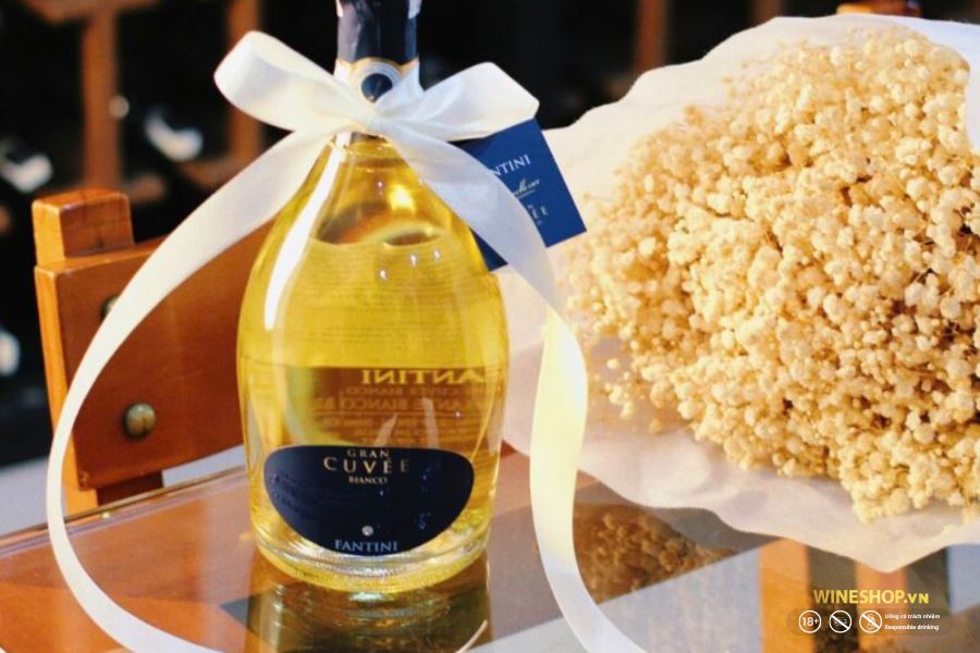 Chai vang nổ Fantini Gran Cuvée Bianco thắt nơ trắng bên bó hoa làm quà tân gia