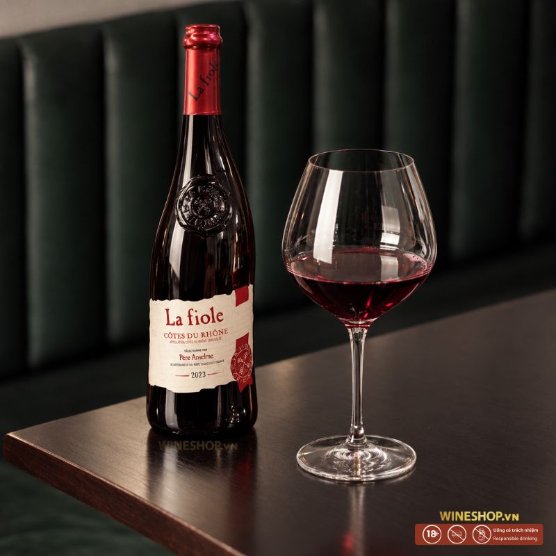 Rượu Vang Pháp La Fiole Côtes Du Rhône Rouge đặt trên bàn gỗ bên cạnh ly rượu đỏ