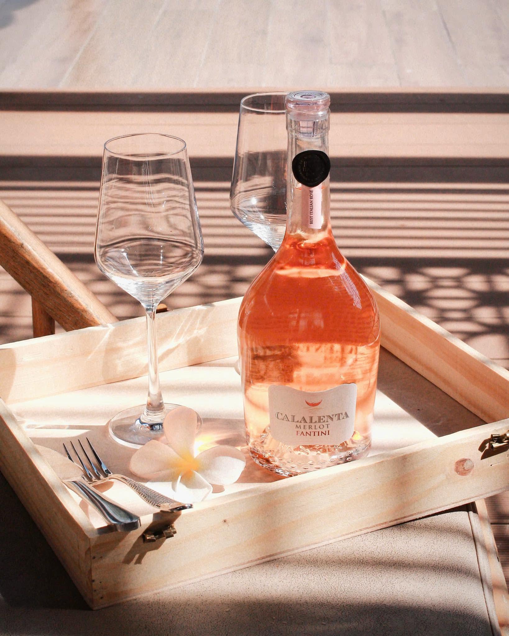 Rượu vang hồng là gì – Chai Fantini Calalenta Merlot Rosé màu hồng đào đặt cạnh ly vang trên khay gỗ dưới ánh nắng tự nhiên.