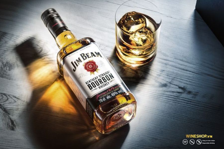 Chai bourbon Jim Beam White bên ly whisky có đá trên mặt bàn gỗ